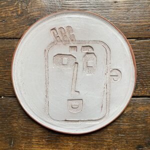 Round, decorative plate with off-white glaze and face made from letterpress block impressions on a wooden floor