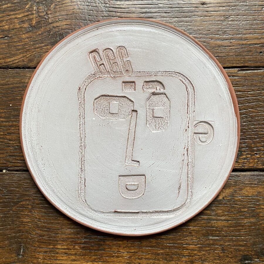 Round, decorative plate with off-white glaze and face made from letterpress block impressions on a wooden floor