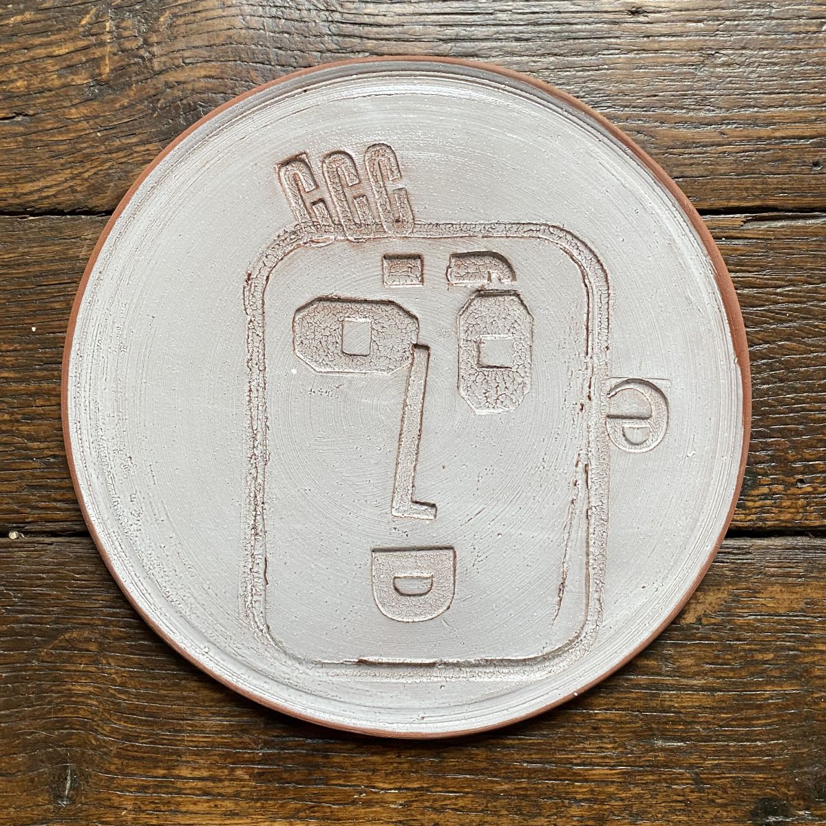 Round, decorative plate with off-white glaze and face made from letterpress block impressions on a wooden floor