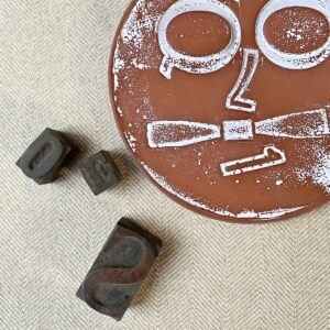 Round, decorative plate with face made from letterpress block impressions on a neutral fabric base