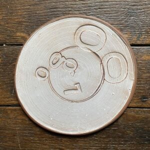 Pale glazed ceramic plate with a face made from impressed letterpress blocks, on wooden floor