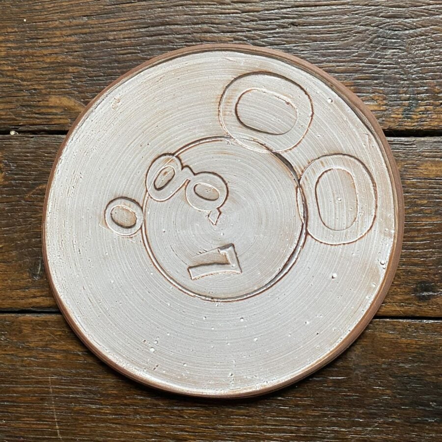 Pale glazed ceramic plate with a face made from impressed letterpress blocks, on wooden floor
