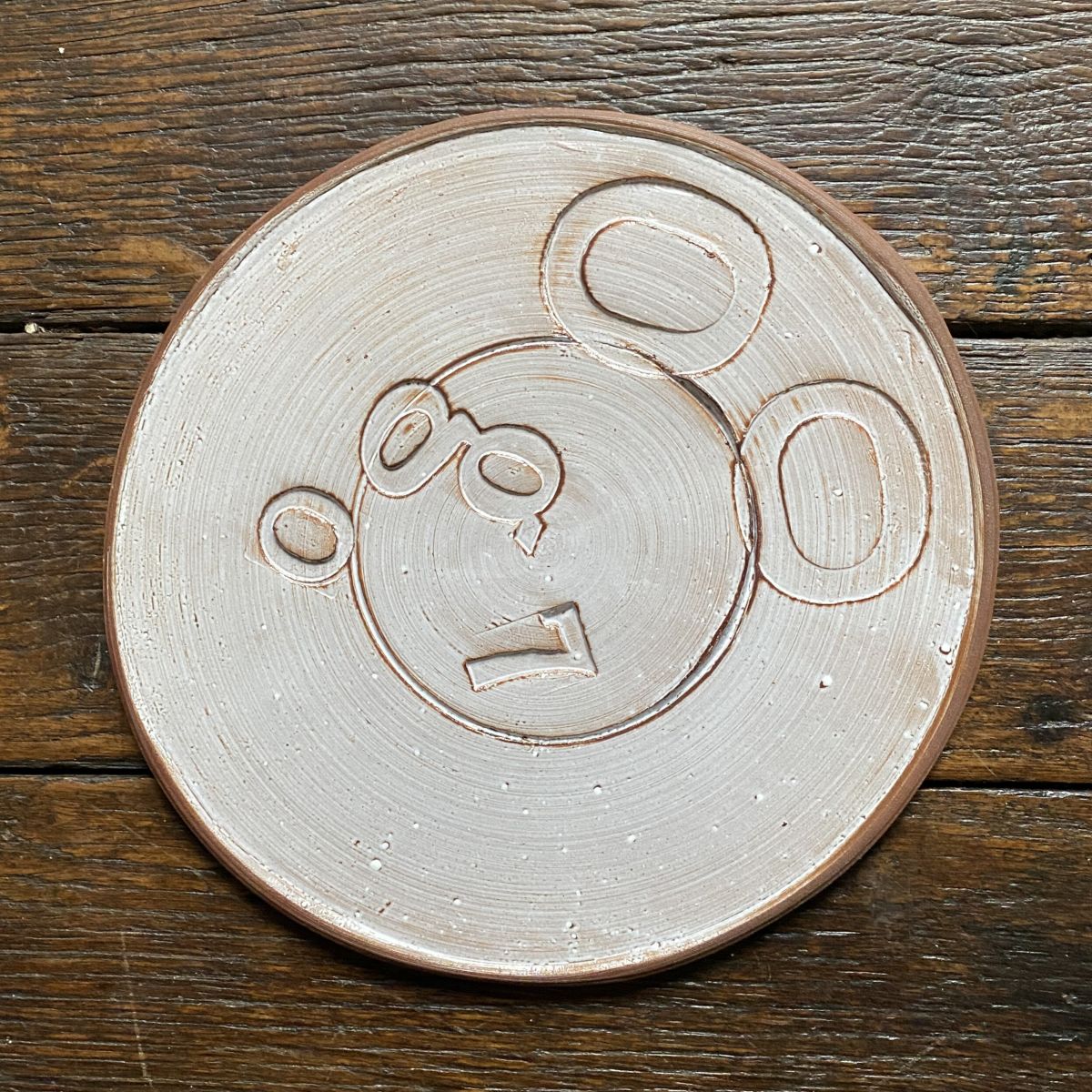 Pale glazed ceramic plate with a face made from impressed letterpress blocks, on wooden floor