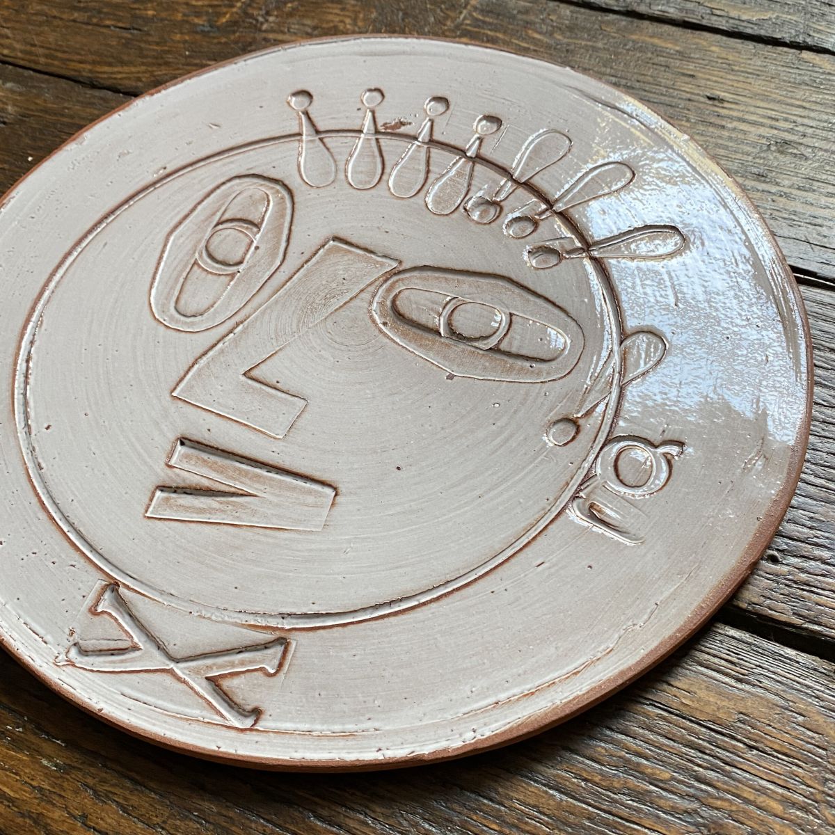 Round, decorative plate with off-white glaze and face made from letterpress block impressions on a wooden floor
