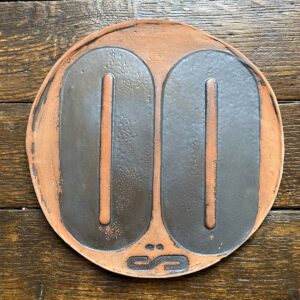 Round, decorative plate with black glaze details and face made from letterpress block impressions on a wooden floor