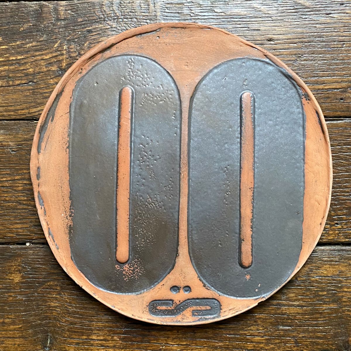 Round, decorative plate with black glaze details and face made from letterpress block impressions on a wooden floor