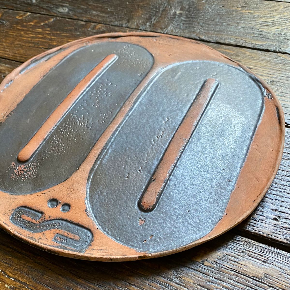 Round, decorative plate with black glaze details and face made from letterpress block impressions on a wooden floor