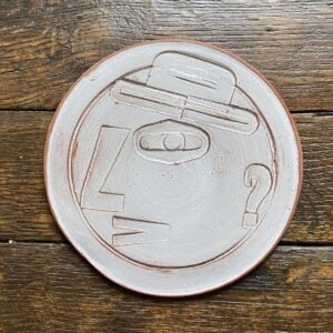Round, decorative plate with off-white glaze and face made from letterpress block impressions on a wooden floor