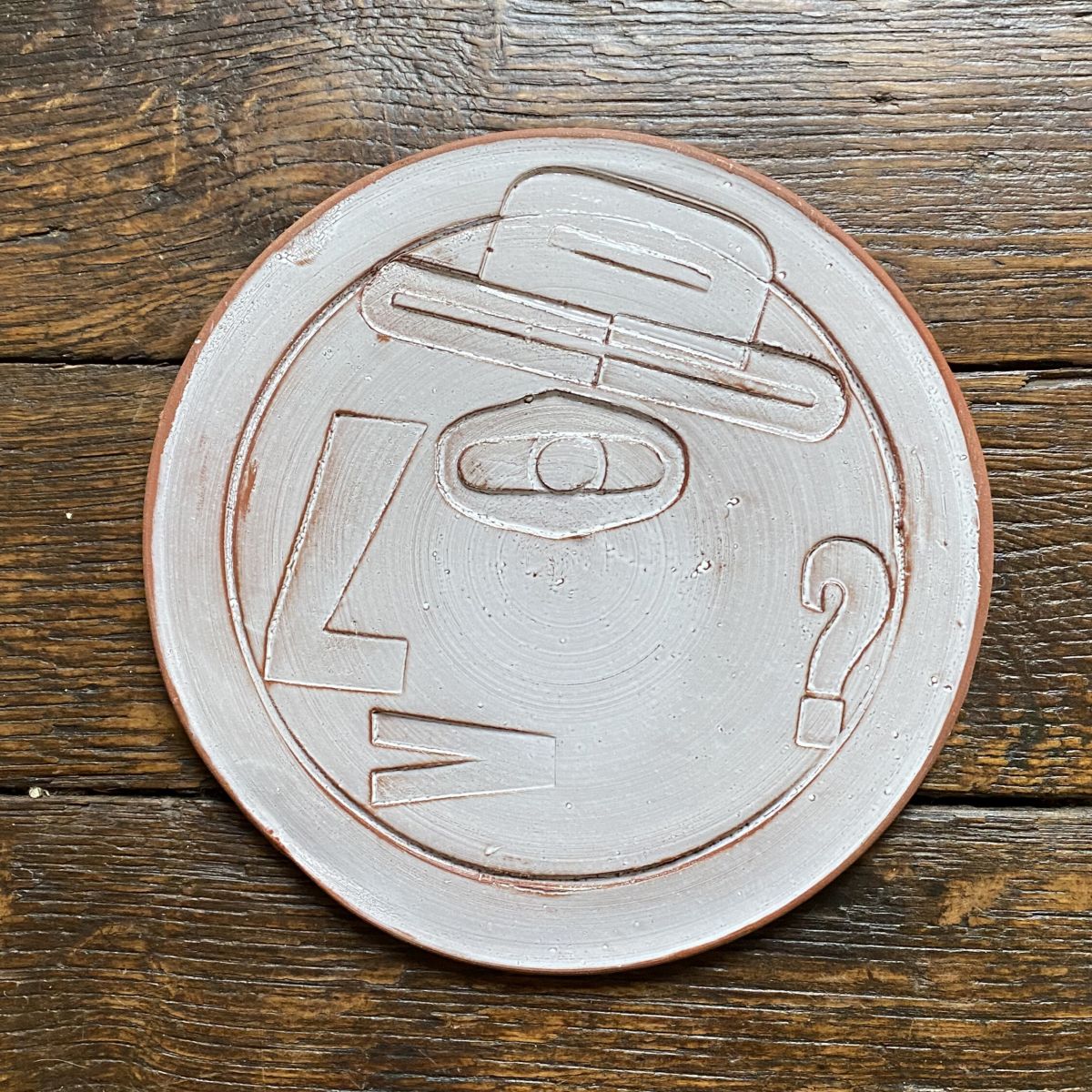 Round, decorative plate with off-white glaze and face made from letterpress block impressions on a wooden floor