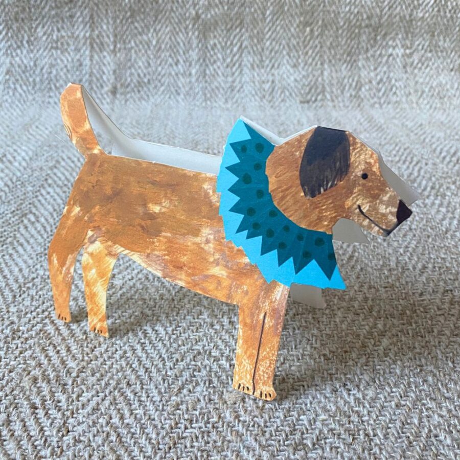 Greetings card in the shape of a brown terrier dog in a blue ruff against a pale fabric background.
