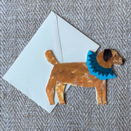 Greetings card in the shape of a brown terrier dog in a blue ruff against a pale fabric background.