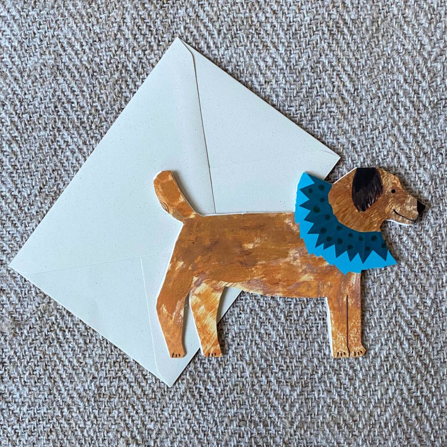 Greetings card in the shape of a brown terrier dog in a blue ruff against a pale fabric background.